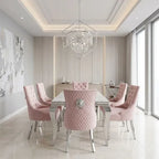 A modern dining room set featuring a rectangular glass-top table surrounded by six tufted pink velvet chairs with silver metal legs and backs, set under a geometric crystal chandelier in a bright, contemporary dining room.