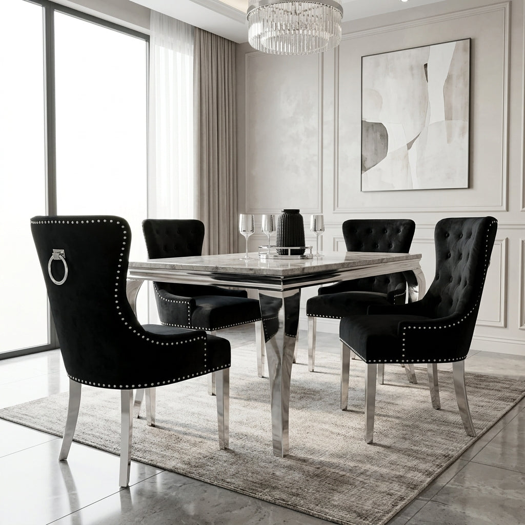 A modern dining room set featuring a rectangular glass-top table with a polished chrome base and six black tufted velvet chairs with silver nailhead trim and ring pulls, arranged on a gray rug in a bright, contemporary room.