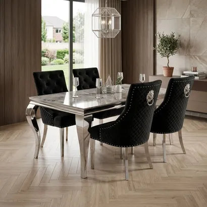 A modern dining room set featuring a rectangular glass-top table with chrome legs and four black upholstered chairs with silver nailhead trim and lion crest backrests, set in a contemporary interior with wood floors and large windows.