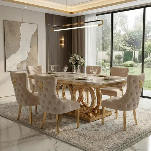 A luxury dining room set featuring a rectangular marble-top dining table with a gold circular metal base, surrounded by six tufted beige velvet chairs with gold accents, set on a patterned area rug in a modern dining room.