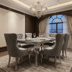 A luxury dining room set featuring a rectangular marble-top dining table with a mirrored silver frame, surrounded by six tufted gray velvet high-back chairs with silver nailhead trim and ring pulls. The room is decorated with a crystal chandelier, beige curtains, and a patterned rug.