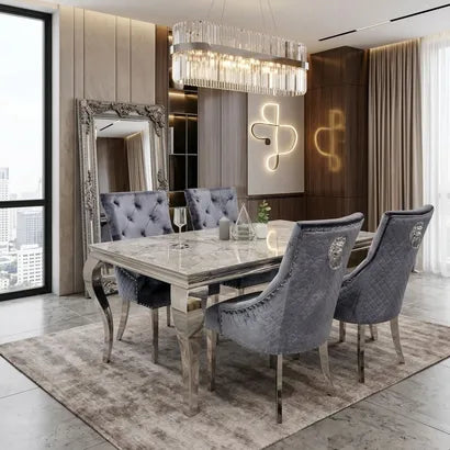 A luxury dining room set featuring a rectangular marble-top dining table with distressed silver legs, surrounded by six tufted gray velvet chairs. The set is styled in a modern, elegant dining room with a crystal chandelier, a large ornate mirror, and warm ambient lighting.