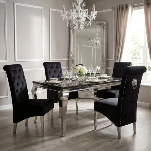 A luxury dining room set featuring a rectangular mirrored glass table with polished silver legs, surrounded by four black tufted velvet dining chairs with silver nailhead trim and silver legs. The room is styled with a large ornate silver mirror, a crystal chandelier, and light gray paneled walls.