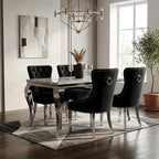 A modern dining room set featuring a rectangular table with a gray marble top and chrome legs, surrounded by four black velvet tufted chairs with silver nailhead trim and ring pulls, set on a geometric patterned rug.