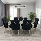A dining room set featuring a rectangular table with a mirrored top and silver legs, surrounded by six black tufted velvet chairs with silver nailhead trim and ring pulls, set in a modern room with gray paneled walls and plants.