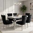 A modern dining room set featuring a rectangular white marble-top table with silver legs and six black velvet baroque-style dining chairs with silver nailhead trim and ring pulls, set in a bright, elegant room with white paneled walls.