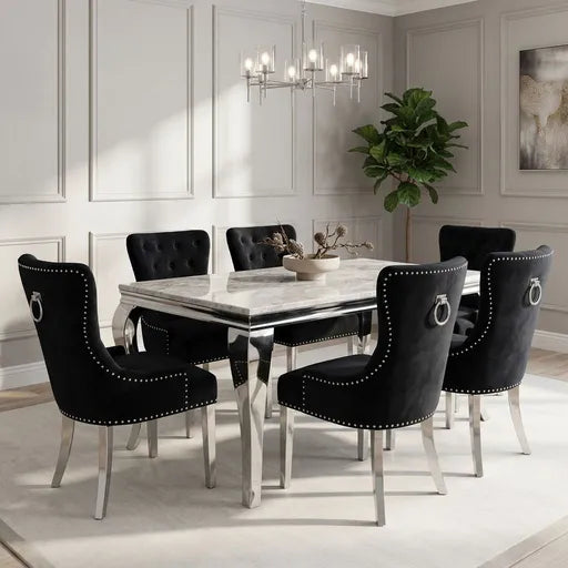 A modern dining room set featuring a rectangular white marble-top table with silver legs and six black velvet baroque-style dining chairs with silver nailhead trim and ring pulls, set in a bright, elegant room with white paneled walls.
