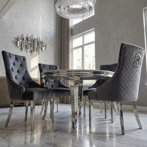 A luxury dining room set featuring a round chrome pedestal table surrounded by four high-back gray upholstered dining chairs with tufted and quilted backrests, studded details, and a lion crest emblem on the back.