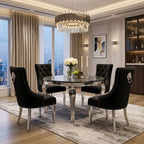 A luxury dining room set featuring a round marble-top table and four black tufted velvet chairs with silver nailhead trim and lion head ring pulls, set on a gray patterned rug in a high-end modern room.
