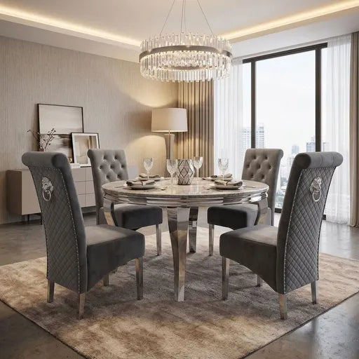 A luxury dining room set featuring a round marble-top table surrounded by four high-back tufted gray velvet chairs with silver metal legs, set on a textured beige rug in a modern, well-lit apartment.
