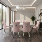 A modern dining room set featuring six blush pink tufted velvet dining chairs with silver lion-head metal accents on the backrests, arranged around a glass-top dining table. The room has a light gray stone accent wall, a large round crystal chandelier, dark wood floors, and tall windows with beige curtains.