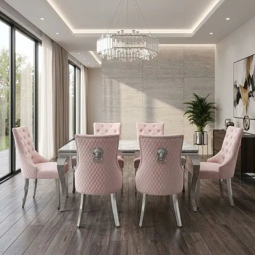 A modern dining room set featuring six blush pink tufted velvet dining chairs with silver lion-head metal accents on the backrests, arranged around a glass-top dining table. The room has a light gray stone accent wall, a large round crystal chandelier, dark wood floors, and tall windows with beige curtains.