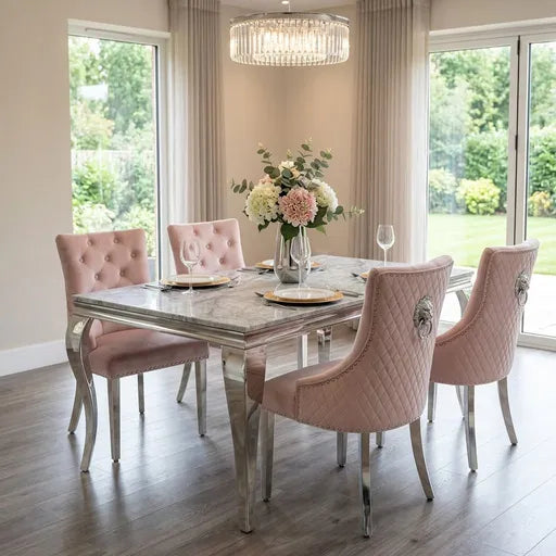 A modern dining room setup featuring a rectangular marble-top dining table surrounded by four blush pink tufted and quilted velvet chairs with silver metal legs. The scene is set in a bright dining room with large windows, beige curtains, a hanging crystal chandelier, and a floral centerpiece on the table.