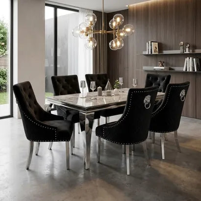 A modern dining room setup featuring a rectangular marble-top dining table surrounded by six black tufted velvet chairs with silver nailhead trim and chrome legs. A multi-light globe chandelier hangs above, set against a background of wood paneling and shelves.