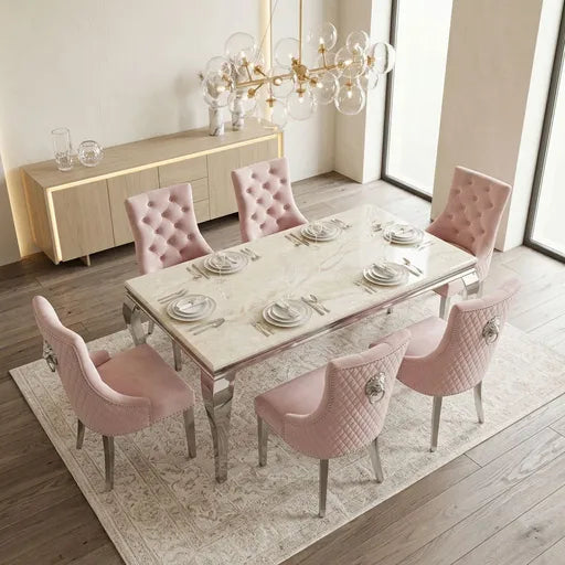 A modern dining room setup featuring a rectangular marble-top dining table surrounded by six blush pink tufted velvet chairs with silver legs, set on a patterned area rug in a bright room with a sideboard and glass chandelier.