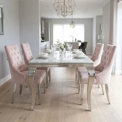 A modern dining room setup featuring a rectangular white marble-top table with a polished silver frame, surrounded by six blush pink tufted velvet dining chairs with silver metal legs.