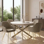 A modern dining set featuring a round white table with a gold-tone X-base and four upholstered chairs in a light taupe velvet, set in a bright, contemporary dining room.