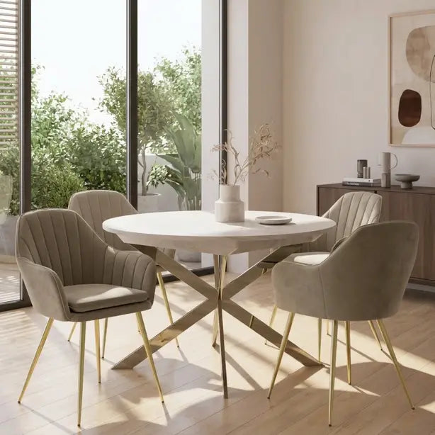 A modern dining set featuring a round white table with a gold-tone X-base and four upholstered chairs in a light taupe velvet, set in a bright, contemporary dining room.