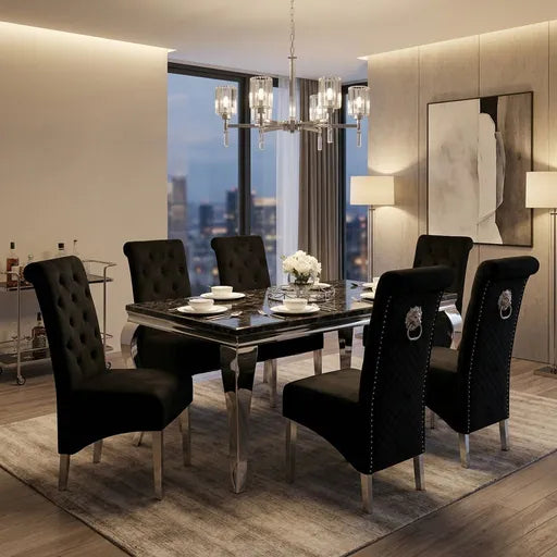A luxury dining room set featuring a rectangular glass-top dining table surrounded by six black tufted velvet chairs with silver metal legs and decorative circular backrests. The set is styled on a patterned gray rug in a high-end modern dining room with a marble wall, city view window, and a gold-toned chandelier overhead.