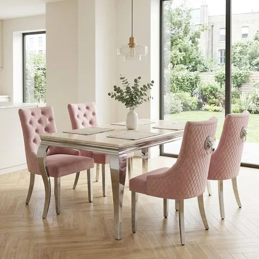 A modern dining set featuring a rectangular table with a light marble top and mirrored legs, surrounded by four pink tufted and quilted velvet chairs in a bright, contemporary dining room.