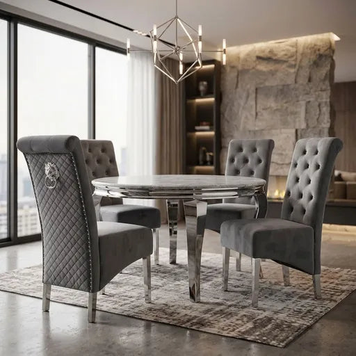 A luxury dining set featuring a round chrome table and four high-back gray tufted velvet chairs with silver legs, styled in a modern dining room with a stone wall and large windows.