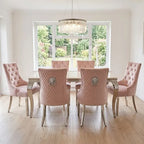 A seven-piece dining set featuring six blush pink tufted velvet chairs with silver lion emblems on the backrests, arranged around a glass-top wooden dining table in a bright, modern dining room.