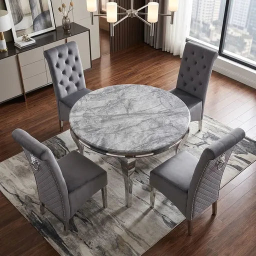 A round dining table with a gray marble-look top and chrome legs, surrounded by four gray upholstered chairs with tufted and quilted backrests, set on a patterned rug in a modern dining room.