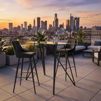Outdoor seating area with high tables and chairs on a rooftop with city skyline in the background during sunset.