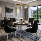 A luxury dining room setup featuring a round chrome-acrylic table surrounded by four black tufted velvet chairs with silver lion head ring pulls. The scene is styled with white plates, crystal glasses, a floral centerpiece, and a large modern chandelier hanging above, set against a gray paneled wall and large windows overlooking a green garden.