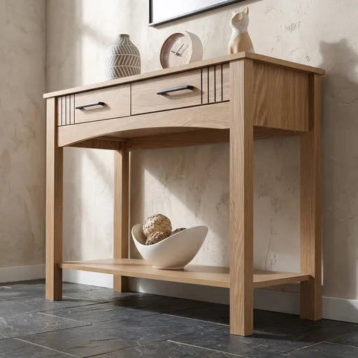 A light natural oak console table with two drawers, decorative slatted details, and an open lower shelf, styled in a minimalist room with a dark slate floor and textured beige walls.