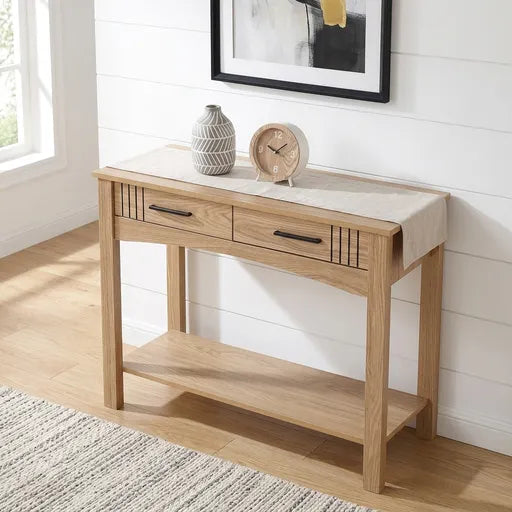 A light oak wood console table with two drawers and an open lower shelf, styled in a bright room with a white shiplap wall, wooden floor, and decorative items on top.