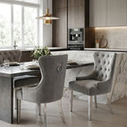 A modern dining room scene featuring two tufted gray velvet dining chairs with silver nailhead trim and metal legs, set around a marble-topped table in a contemporary kitchen interior.