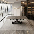 A modern rectangular dining table with a dark gray stone-look top and sculptural black metal base, shown in a luxury contemporary dining room with large windows, a wine cellar, and a hanging brass chandelier.