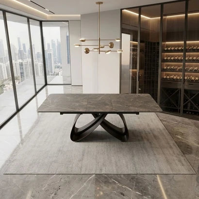 A modern rectangular dining table with a dark gray stone-look top and sculptural black metal base, shown in a luxury contemporary dining room with large windows, a wine cellar, and a hanging brass chandelier.