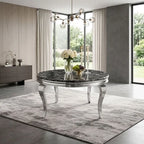 A round dining table with a black marble-patterned top and curved silver legs, styled in a modern dining room with a gray rug, curtains, and a glass vase of flowers on top.