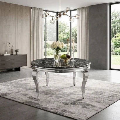 A round dining table with a black marble-patterned top and curved silver legs, styled in a modern dining room with a gray rug, curtains, and a glass vase of flowers on top.