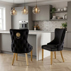 Two black upholstered dining chairs with gold-tone legs and hardware, shown in a modern kitchen setting. One chair features a tufted back with a lion head ring pull, and the other has a quilted back with nailhead trim.