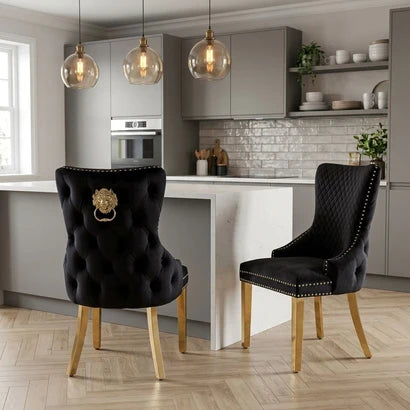 Two black upholstered dining chairs with gold-tone legs and hardware, shown in a modern kitchen setting. One chair features a tufted back with a lion head ring pull, and the other has a quilted back with nailhead trim.