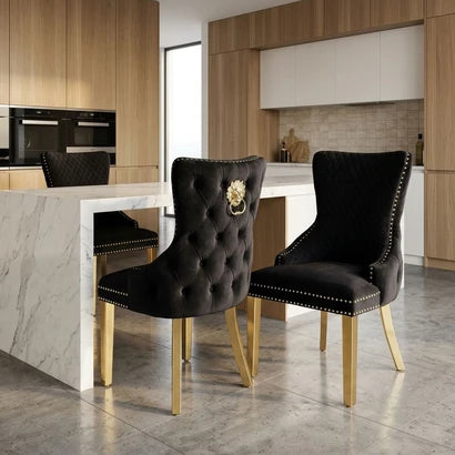 Two black velvet dining chairs with gold legs and nailhead trim, shown in a modern kitchen setting. One chair features a tufted back with a gold lion emblem.