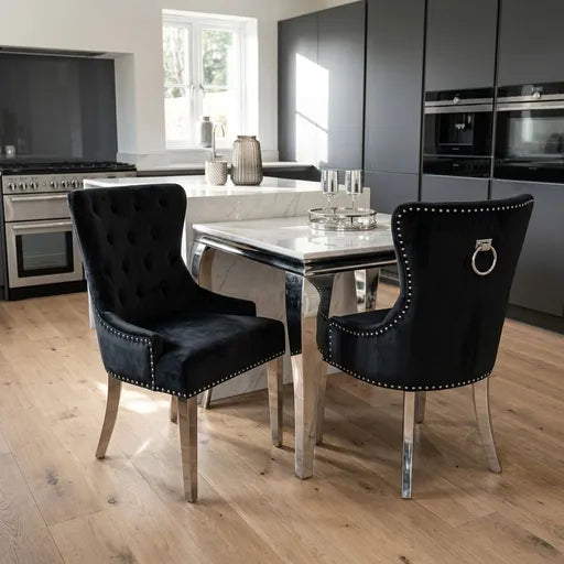 Two black velvet dining chairs with silver nailhead trim and ring pulls, positioned around a marble-top dining table in a modern kitchen setting with light wood floors.
