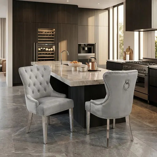 Two  gray tufted dining chairs with silver nailhead trim and metal legs, positioned at a kitchen island in a modern kitchen setting.