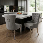 Two gray tufted dining chairs with silver nailhead trim and ring pulls, positioned at a kitchen island in a modern kitchen setting with wood floors and dark cabinetry.