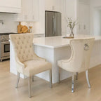 Two tufted cream dining chairs with silver metal legs and nailhead trim, positioned in a bright, modern kitchen with white cabinetry and a white island.