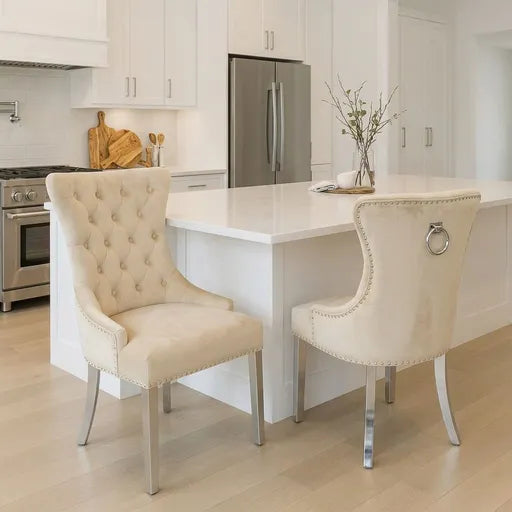 Two tufted cream dining chairs with silver metal legs and nailhead trim, positioned in a bright, modern kitchen with white cabinetry and a white island.