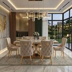 A luxury dining room featuring a rectangular light wood dining table surrounded by six tufted champagne-colored upholstered chairs with gold lion head accents. The room is decorated with a modern geometric chandelier, a patterned area rug, and large floor-to-ceiling windows overlooking a garden patio at dusk.