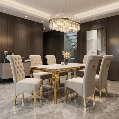 A luxury dining room set featuring a rectangular white marble dining table with a gold frame and six matching tufted beige upholstered chairs with gold legs, under a large crystal chandelier.