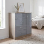 A modern 5-drawer chest of drawers in a gray and light wood finish, styled in a bright bedroom next to a window with a framed print and vase on top.