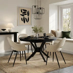 A modern dining room setup featuring a round black table with a central pedestal base, surrounded by four cream-colored upholstered chairs with black legs. The scene is styled with a woven jute rug, a black side table with a lamp, and a window with a built-in bench seat in the background.