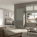 A tall, modern two-door wardrobe in a matte gray finish stands in a contemporary, sunlit living and sleeping room. The room features neutral beige and gray tones, with a bed, sofa, and a wooden coffee table visible in the background near a large window.