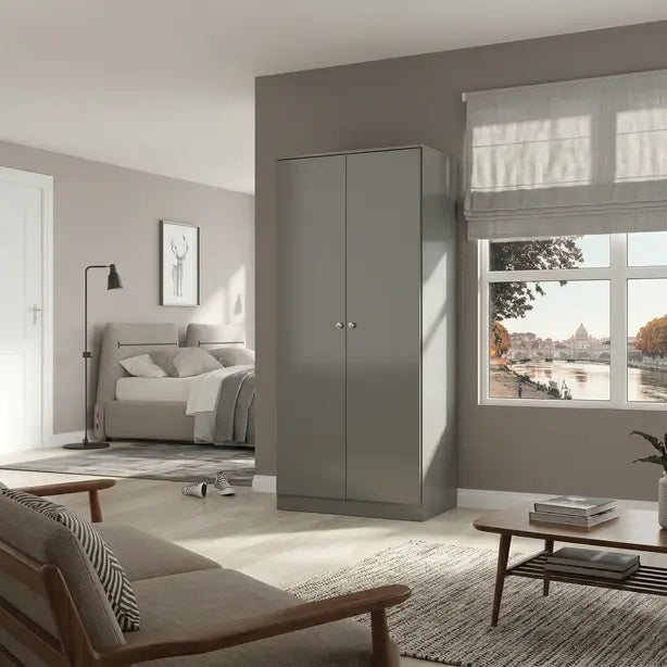 A tall, modern two-door wardrobe in a matte gray finish stands in a contemporary, sunlit living and sleeping room. The room features neutral beige and gray tones, with a bed, sofa, and a wooden coffee table visible in the background near a large window.